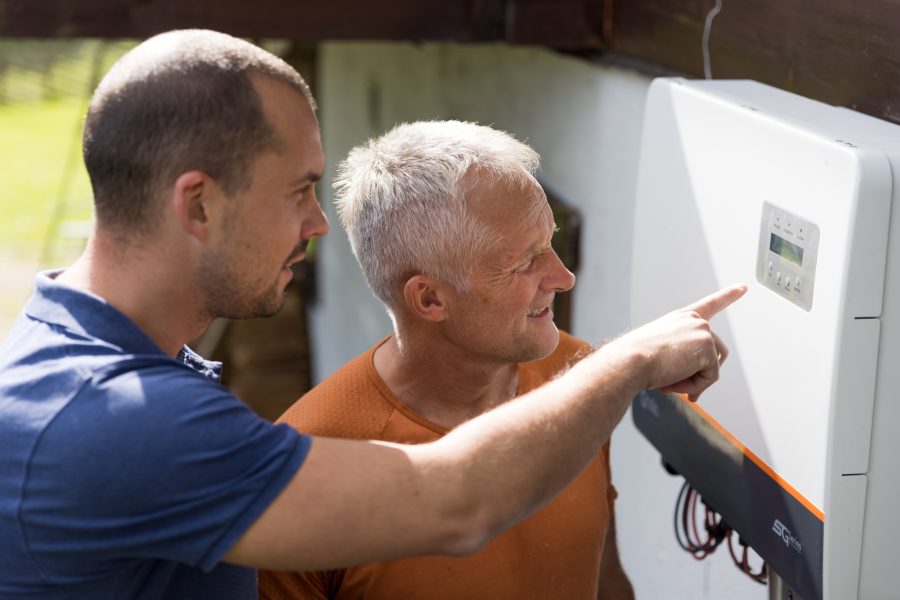 Elektriker i blå t-skjorte og mann i oransje skjorte inspiserer en vekselsretter montert på husvegg.