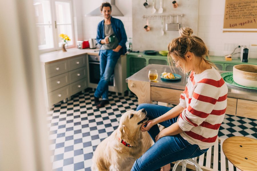 en mann med en kaffekopp i hånda og en smilende dame i hvit øg rød stripete genser som sitter på en stol på kjøkkenet og koser med en golden retriver.