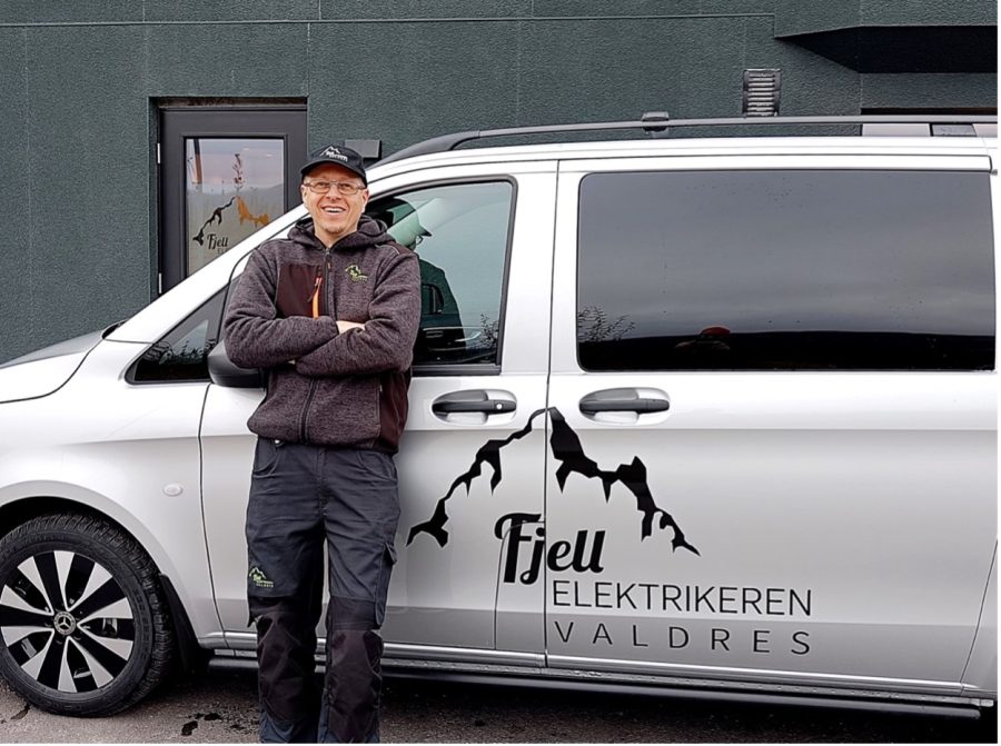 Mannlig elektriker i sort arbeidstøy og caps står smilende foran sølvgrå varebil med logo: fjelle elektrikeren valdres