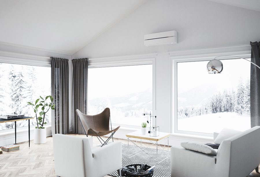 hvit varmepumpe som er montert på en hvit vegg over to store vinduer, og en grå sofa og to stoler i forgrunnen med en grønn plante og snølandskap utenfor boligen.