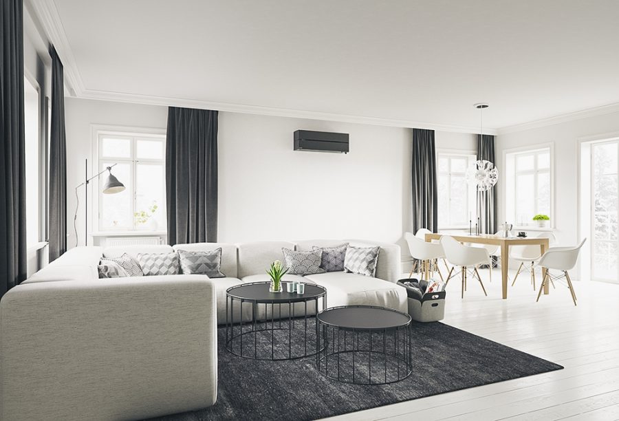 sort varmepumpe på som er montert på en hvit vegg i en stue med sort gardiner, grå hjørnesofa med sort teppe og to runde sorte bord, og en spisestue med trebord og hvite stoler til høyre i bakgrunnen og hvitt tregulv.